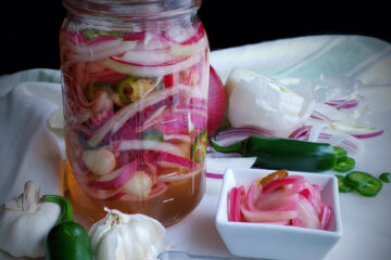 An overhead image of these Quick and Easy Pickled Onions shows a large mason jar full of crisp onions, thinly sliced jalapeno peppers and garlic. The sweet smell of the apple cider vinegar and sugar practically comes through the image.