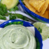A closeup shot of this Super Simple 5-Minute Avocado Buttermilk Dressing. This dressing looks so light and flavorful with its creamy texture and fresh herbs. In the background is a bowl of crisp tortilla chips, more fresh herbs, and avocado and lime that have been cut in half. They are at their peak freshness.