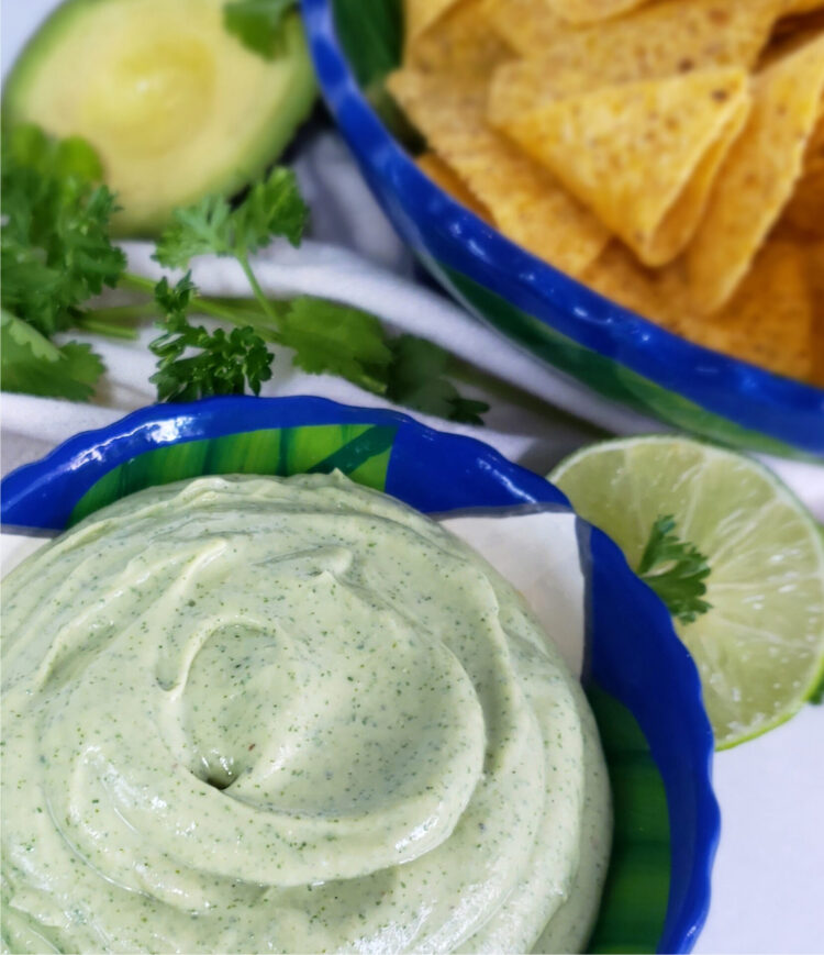 A closeup shot of this Super Simple 5-Minute Avocado Buttermilk Dressing. This dressing looks so light and flavorful with its creamy texture and fresh herbs. In the background is a bowl of crisp tortilla chips, more fresh herbs, and avocado and lime that have been cut in half. They are at their peak freshness.