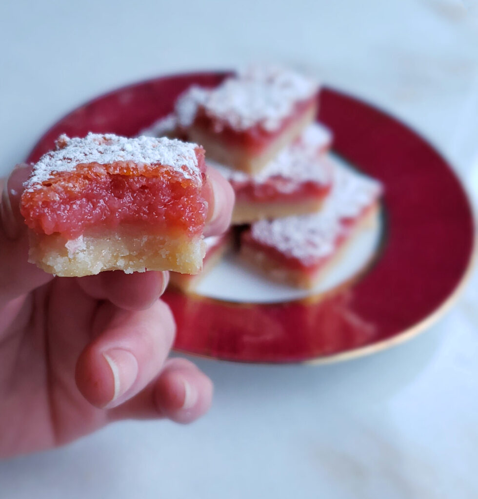 A close up shot of a Tempting 2 Bite Blood Orange Bar with a bite playfully taken out of it. The shortbread crust shows flecks of coconut. Blood Orange zest is sprinkled throughout the filling and the top of the bar is dusted with powdered sugar.