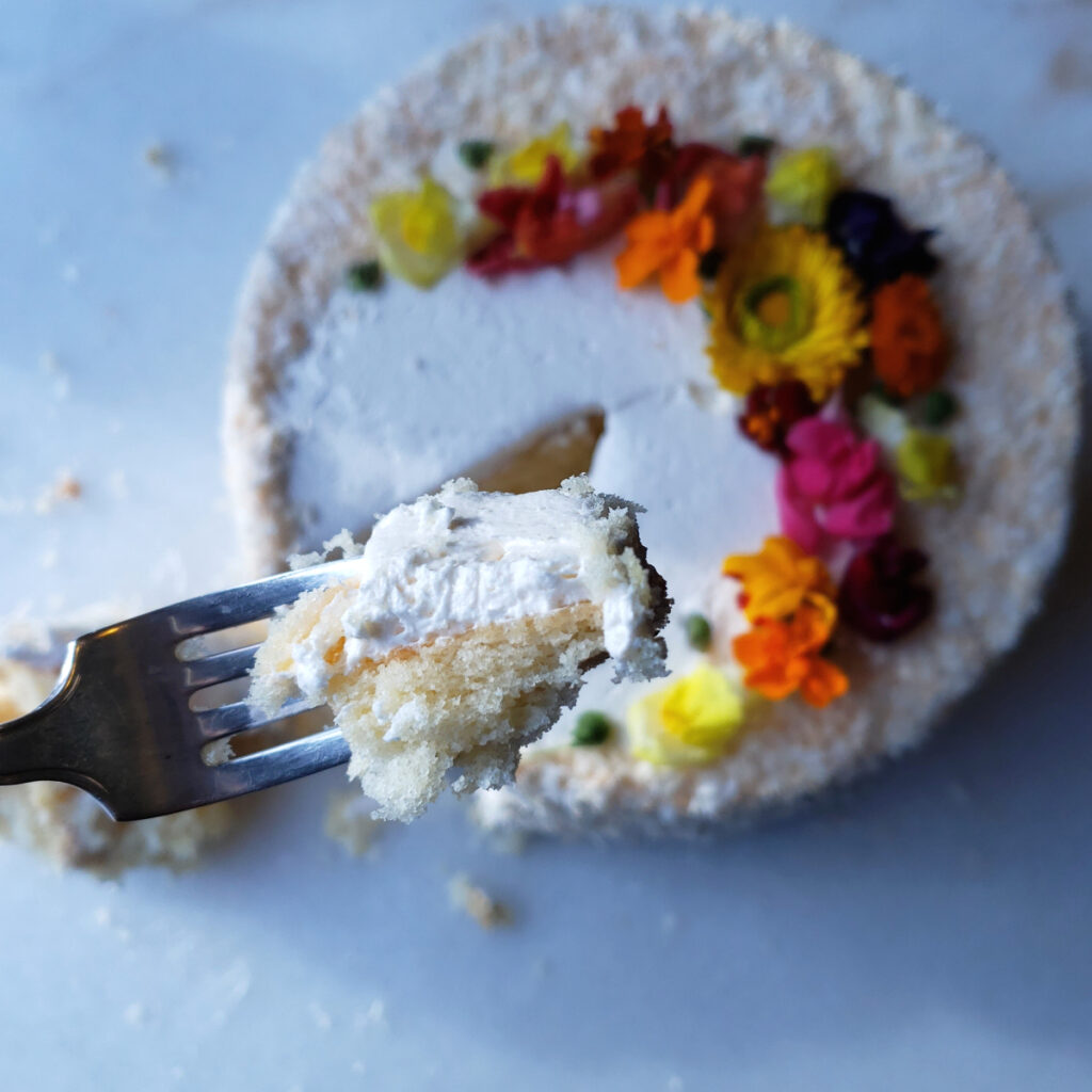 Fresh edible flowers and lightly toasted coconut adorn this Easy Pineapple Coconut Cake with 5-Ingredient Rum Buttercream. A fork is lifted up to reveal the cake's moist, delicate crumb and light buttercream.