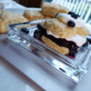 A closeup shot of a Super Tender 8-Ingredient Lemon Shortcake sits at the center of a plate. A warm blueberry compote and fresh whipped cream is sandwiched between two halves of a shortcake. A dalop of whipped cream, fresh lemon zest and a few extra fresh berries top the towering treat.