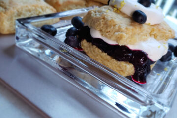 A closeup shot of a Super Tender 8-Ingredient Lemon Shortcake sits at the center of a plate. A warm blueberry compote and fresh whipped cream is sandwiched between two halves of a shortcake. A dalop of whipped cream, fresh lemon zest and a few extra fresh berries top the towering treat.