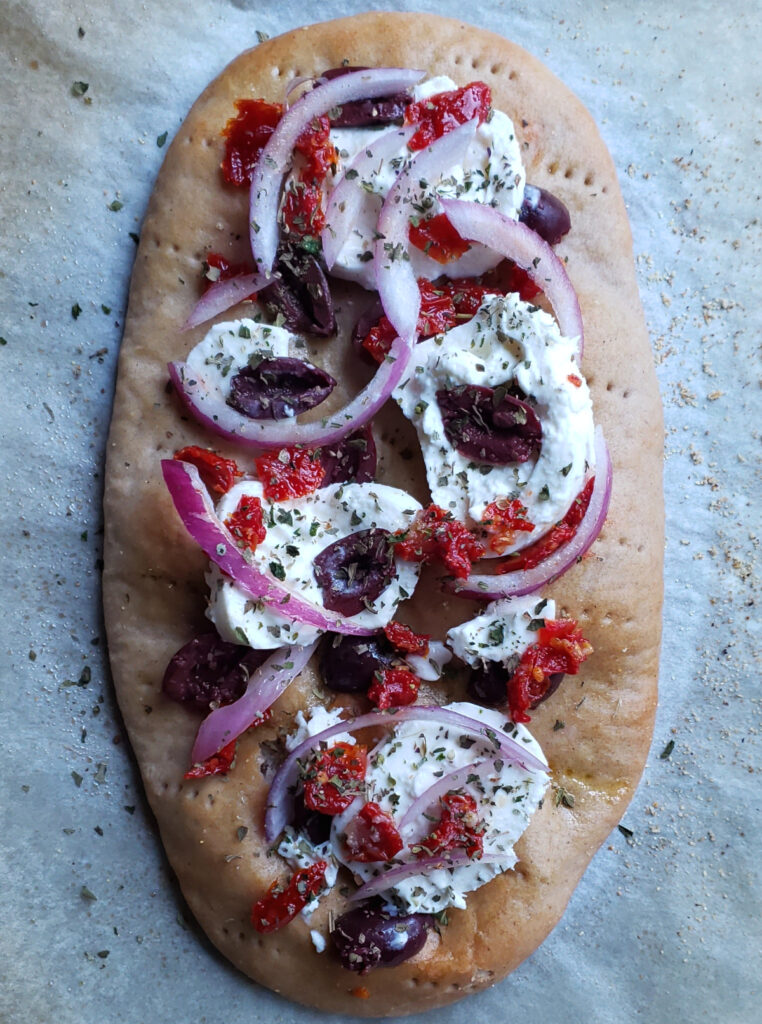 A Mediterranean style pizza featuring this Easy 7_Ingredient Blue Corm Flatbread stands ready to go in the oven. Topped with creamy burrata cheese, kalamata olives, sun dried tomatoes, red onions and dried herbs.