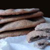 A beautiful stack of fresh bakes Easy 7-Ingredient Rustic Buckwheat Flatbreads rest upon a sheet of parchment paper. One of the flatbreads is torn in half to reveal its light texture inside.