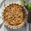A beautifully baked Sour Cream Dutch Apple Pie with Easy Rye Crust sits atop a cooling rack in the warm sunlight. A fresh Granny Smith Apple is cut in half in the top right corner of the frame. A crisp rye crust, golden crumb topping with oats and pecans adorn the pie. The smell of cinnamon, nutmeg and brown sugar practically rise from the picture.