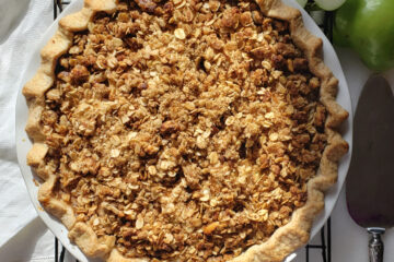 A beautifully baked Sour Cream Dutch Apple Pie with Easy Rye Crust sits atop a cooling rack in the warm sunlight. A fresh Granny Smith Apple is cut in half in the top right corner of the frame. A crisp rye crust, golden crumb topping with oats and pecans adorn the pie. The smell of cinnamon, nutmeg and brown sugar practically rise from the picture.