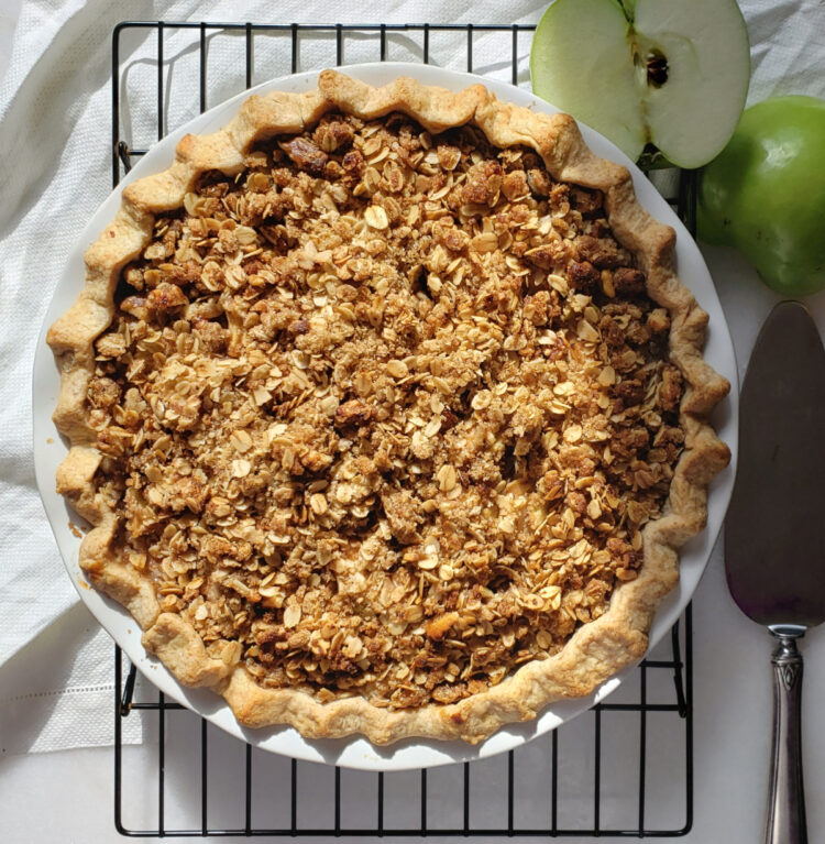 A beautifully baked Sour Cream Dutch Apple Pie with Easy Rye Crust sits atop a cooling rack in the warm sunlight. A fresh Granny Smith Apple is cut in half in the top right corner of the frame. A crisp rye crust, golden crumb topping with oats and pecans adorn the pie. The smell of cinnamon, nutmeg and brown sugar practically rise from the picture.