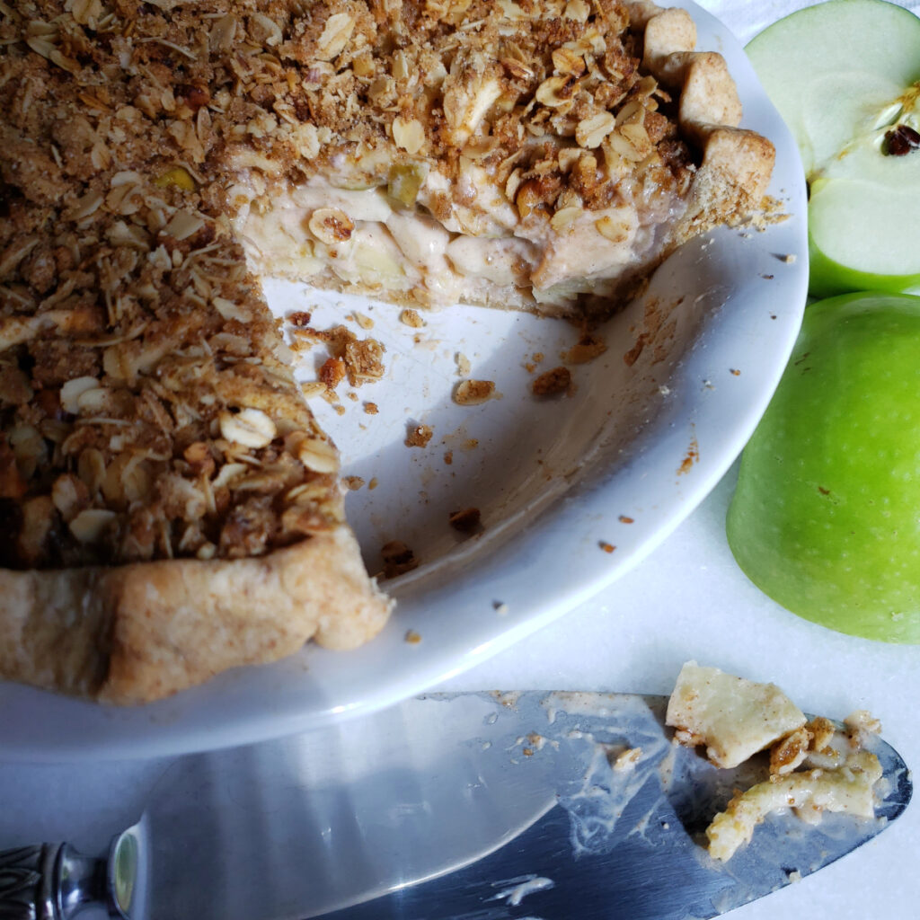A beautiful slice of Sour Cream Dutch Apple Pie with Easy 3-Step Rye Crust sits in the middle of a dessert plate. A fork is lifted to the center of the frame to reveal the creamy apple filling, crisp rye crust and hearty crumb topping.
