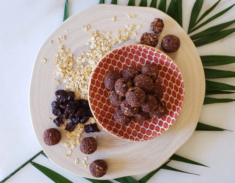 A plate of 7-Ingredient No Bake Energy Bites are stacked in a perfect pyramid. Oats and dried cherries are strewn across the wood cutting board on which the plate sits.