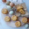 On top of a rustic, wooden board rests fresh Bold Ginger Mango Muffins with Sesame Streusel. Fresh farm eggs and are pictured in the background.