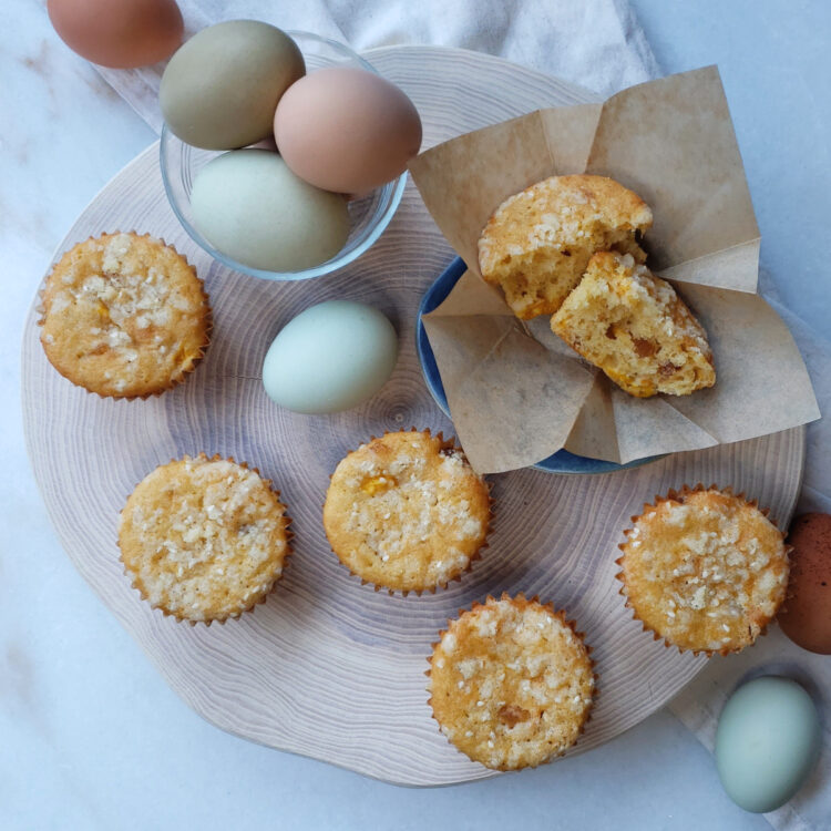 On top of a rustic, wooden board rests fresh Bold Ginger Mango Muffins with Sesame Streusel. Fresh farm eggs and are pictured in the background.