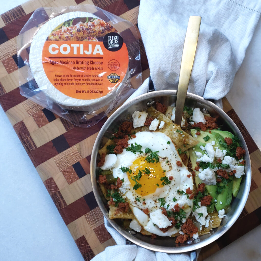 A beautiful single serving of Easy 6-Ingredient Authentic Chilaquiles sits atop a decorative, wood cutting board. A round of fresh Cotija cheese sits off to the side. Toothy tortilla chips tossed in salsa verder are topped with a sunny side up egg, slices of fresh avocado, chorizo crumbles and a generous topping of Rizo Bros Cotija cheese.