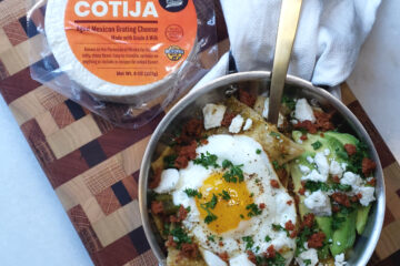 A beautiful single serving of Easy 6-Ingredient Authentic Chilaquiles sits atop a decorative, wood cutting board. A round of fresh Cotija cheese sits off to the side. Toothy tortilla chips tossed in salsa verder are topped with a sunny side up egg, slices of fresh avocado, chorizo crumbles and a generous topping of Rizo Bros Cotija cheese.