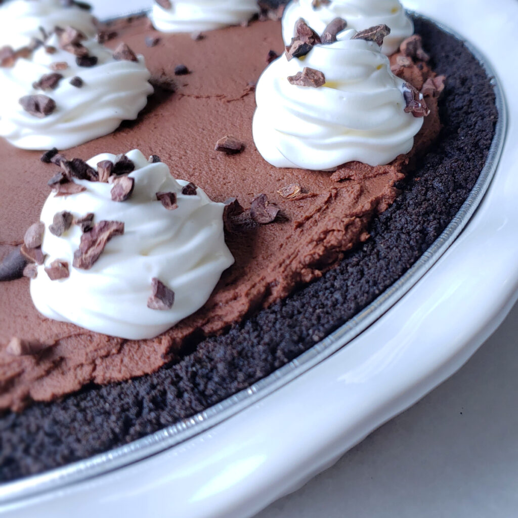 A closeup shot of this Decadent 5-Ingredient Whipped Ganache Pie. A crisp chocolate cookie crust, rich, whipped dark chocolate ganache is finished with a dallop of sweetened whipped cream and a sprinkle of cocoa nibs.