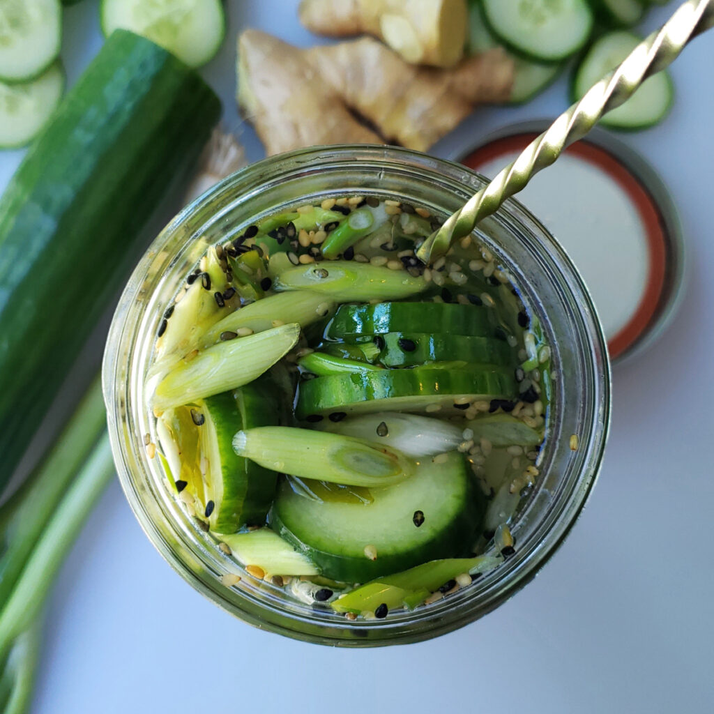 A beautiful jar of Easy 10-Minute Ginger Sesame Pickles sits pretty in the center of a cutting board - surrounded by fresh cucumbers, scallions and ginger.