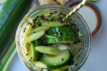 A beautiful jar of Easy 10-Minute Ginger Sesame Pickles sits pretty in the center of a cutting board - surrounded by fresh cucumbers, scallions and ginger.