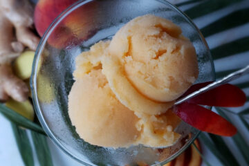 An elegant, frosty glass of this Heavenly 3-Ingredient Peach Ginger Kombucha Sorbet sits nestled among fresh peaches and sliced ginger root. There is a warm tropical vibe to the picture.