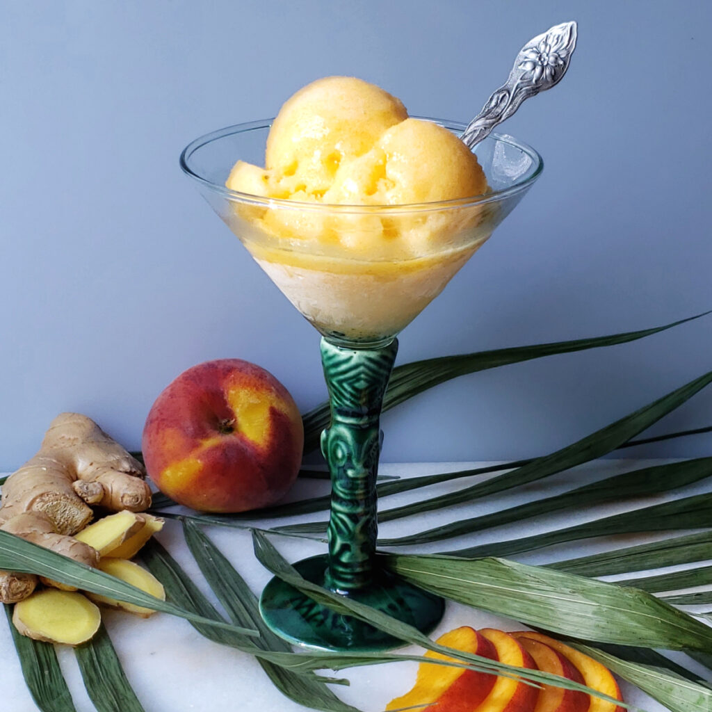 A tall glass of Heavenly 3-Ingredient Peach Ginger Kombucha Sorbet is pictured among fresh peaches and ginger root slices.