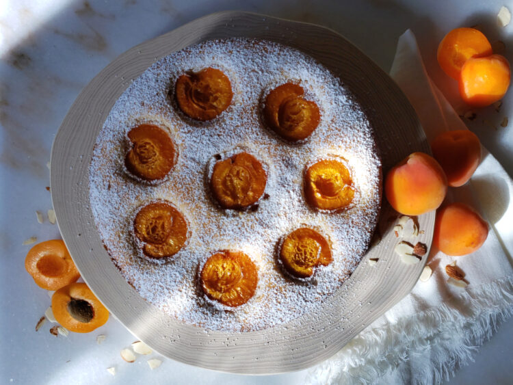 A whole Simply Delicious 8-Star Almond Cake rests upon a cake stand in the sunlight. Fresh apricots and almonds adorn the surface of the counter.