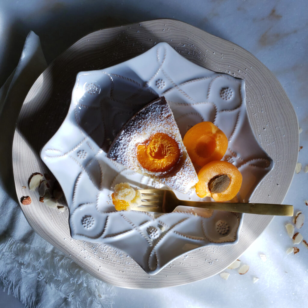 A slice of this Simply Delicious 8-Star Almond Cake sits atop a small dessert plate. A single bite is perched at the tip of a fork. Sunlight dances across the top of the cake and illuminates the apricots.