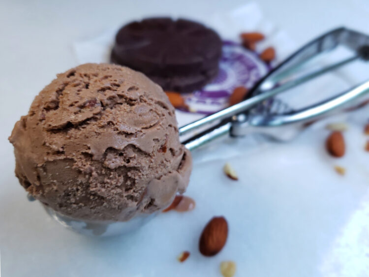 A cool and refreshing scoop of Easy 7-Ingredient Mexican Chocolate Ice Cream is nested in an ice cream scoop. Disks of Mexican Chocolate and toasted almonds are in the background.