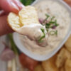 A deliciously dippable bowl of Super Simple 15 Minute Onion Dip is nested among a platter of golden, kettle cooked potato chips. Fresh green onions and shallots adorn the platter. A closeup shot of a chip that has just been dipped.