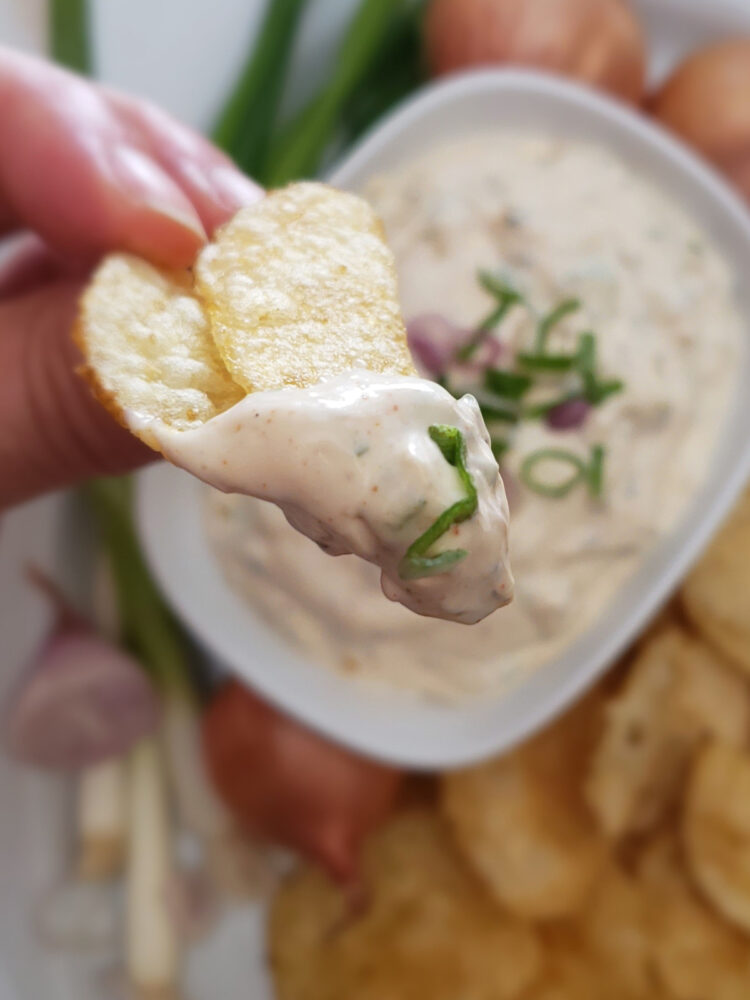 A deliciously dippable bowl of Super Simple 15 Minute Onion Dip is nested among a platter of golden, kettle cooked potato chips. Fresh green onions and shallots adorn the platter. A closeup shot of a chip that has just been dipped.