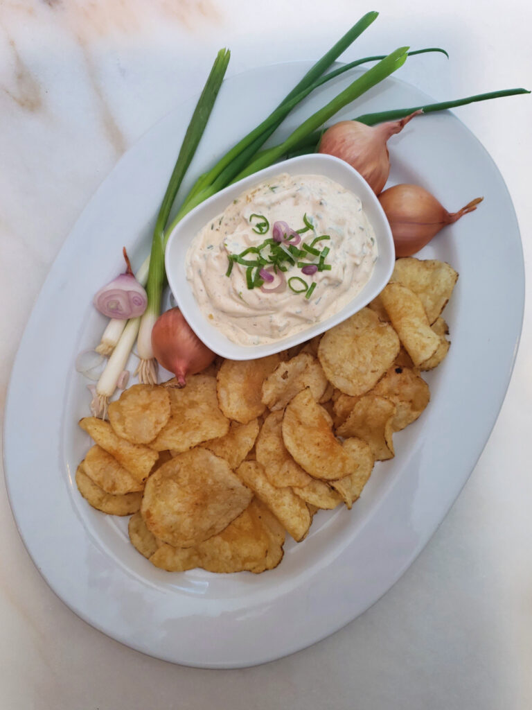 A deliciously dippable bowl of Super Simple 15 Minute Onion Dip is nested among a platter of golden, kettle cooked potato chips. Fresh green onions and shallots adorn the platter.