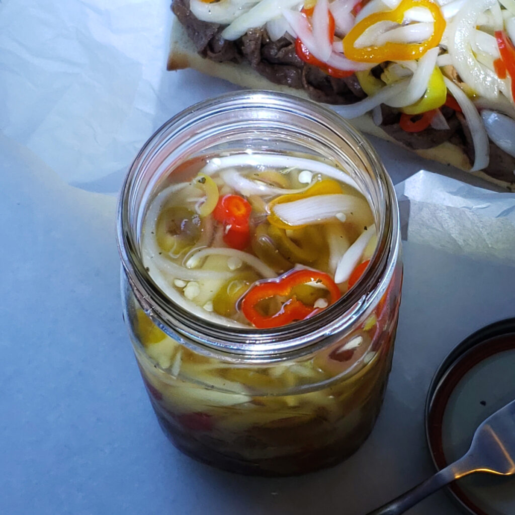 A jar of Giardiniera sits in the forefront. In the background, a shaved steak sandwich on a french roll is generously topped with this Simple Sweet and Spicy 15-Minute Giardiniera