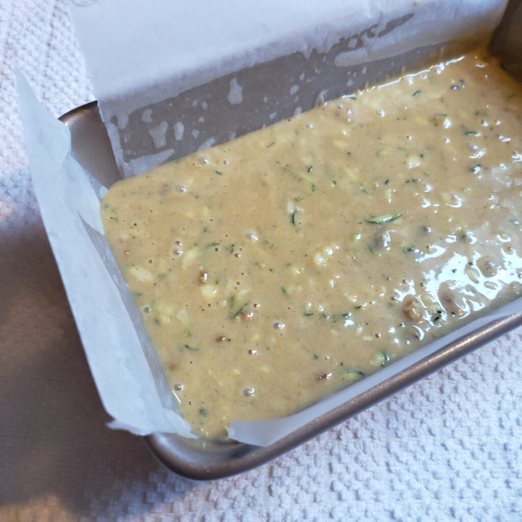 A baking pan is prepared and filled with the batter for this Best Old Fashioned Zucchini Bread in 60 Minutes. The pan is lined with parchment paper to prevent sticking. 