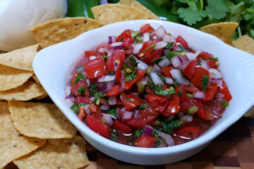 A bowl of Fresh Pico do Gallo in 10 Easy Minutes is surrounded by rustic corn tortilla chips, jalapenos, cilantro, onions, tomatoes and limes.