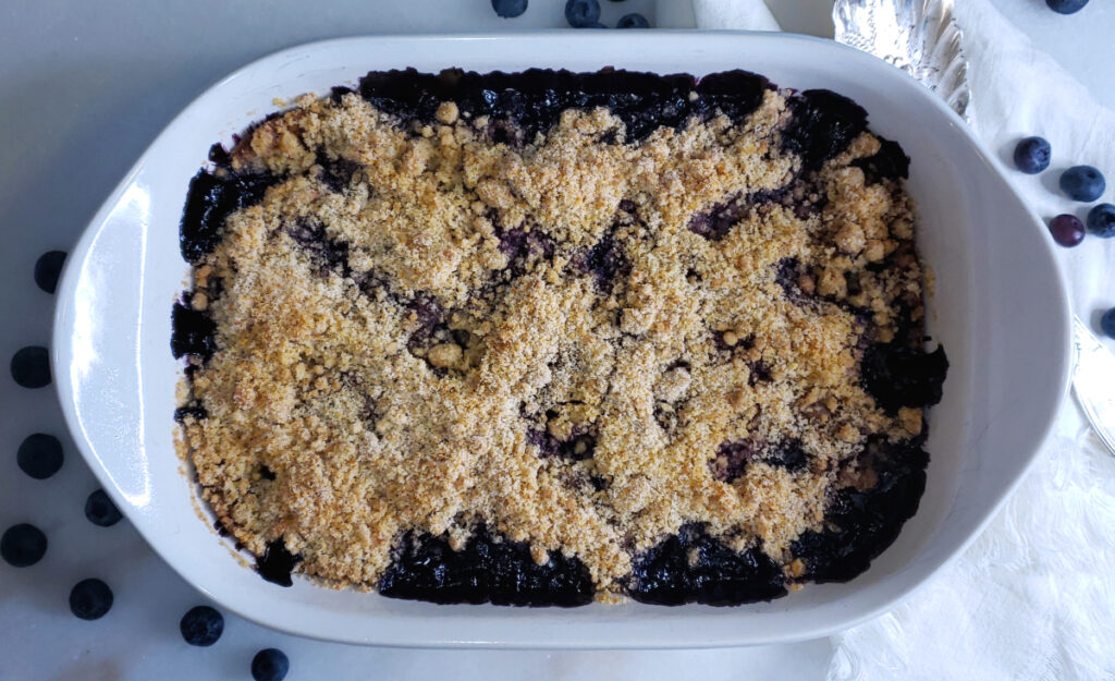 Hot out of the oven, a baking dish of bubbly hot Amazing Honolulu Blueberry Crisp in 90 Minutes sits cooling on a white marble countertop.