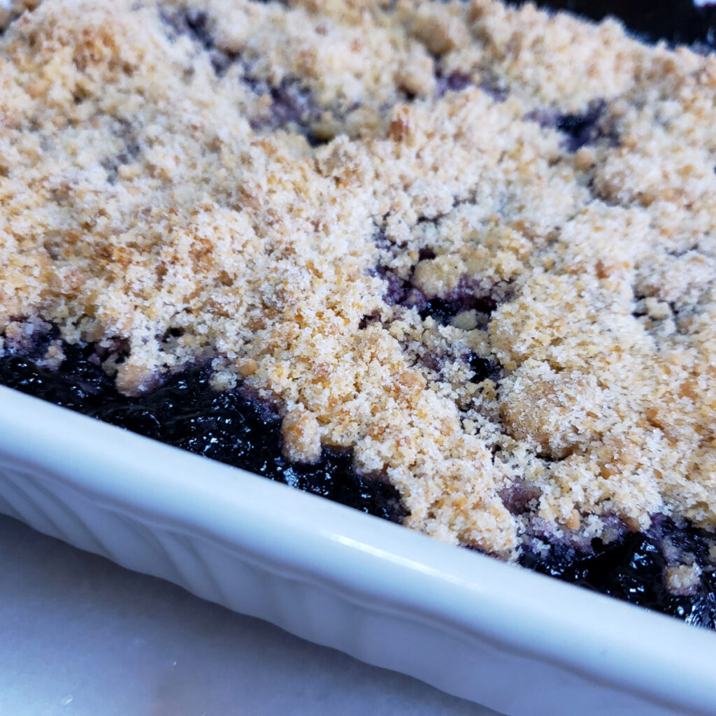  Hot out of the oven, a baking dish of bubbly hot Amazing Honolulu Blueberry Crisp in 90 Minutes sits cooling on a white marble countertop.