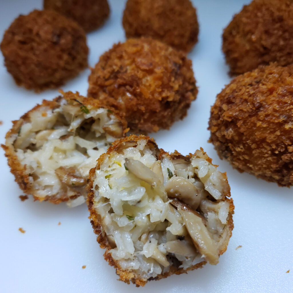 A closeup of a crispy, golden brown Easy Mushroom Arancini in 2 Hours. The inside is broken open to reveal its filling of mushrooms, herbs, and a pearl of mozzarella.