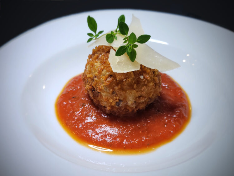 A pristine plate is dresses with a rich tomato sauce. A single mushroom arancini sits in the middle of the sauce and is finished with a few shavings of fresh parmesan cheese and fresh herbs as the feature image for Easy Mushroom Arancini in 2 Hours.