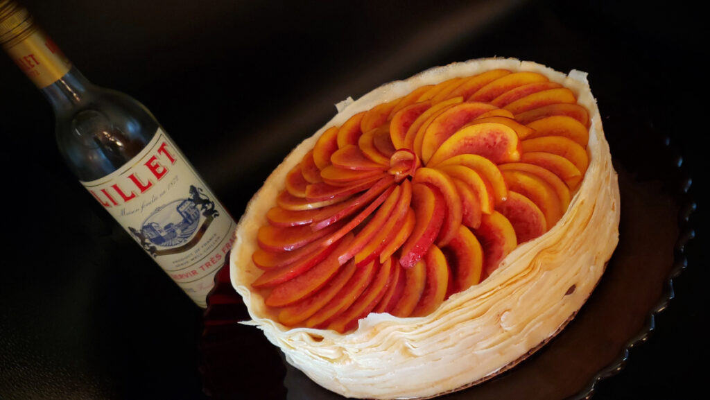 A bottle of Lillet Rosé is featured in the background of this BEst 20 Layer Cannoli Cream Crepe Cake. A beautiful crepe cake topped with fresh nectarines sits in the forefront waiting to be cut into.