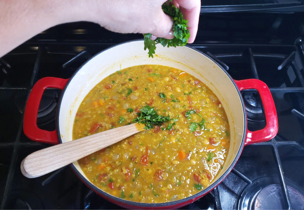 A finished pot of this beautiful Easy Vegetarian Curried Lentil Soup in 1 Hour is featured.  A hand hovers over the top and is sprinkling in the fresh parsley at the very end of the cooking process.