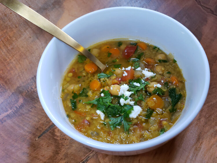 A simple, white porcelain bowl sits atop a natural wood table and reveals a generous portion of this Easy Vegetarian Curried Lentil Soup in 1 Hour. A light drizzle of olive oil, feta crumbles and fresh parsley tops the bowl. A spoon is poised to take a first taste of this hearty and healthy soup.