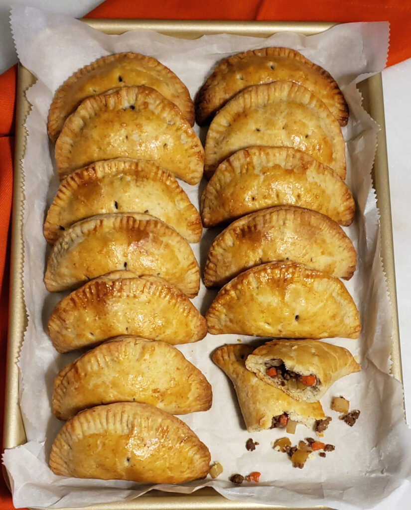 A tray of perfectly shaped, hot-out-of-the-oven Easy Opening Day Michigan Pasties. These perfect little half-moon hand pies are lined up in two rows. The crust is glazed with an egg wash and is a rich golden brown. One pasty is split in half, revealing a delicious filling of meat or plant protein, onions, potatoes, carrots and rutabaga.