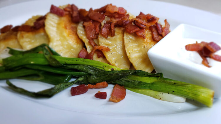 A tray of Vibrant 2-Color Sweet Potato Pierogi sit upon a bed of grilled green onions. Crisp bacon crumbles top the pierogi. A dish of sour cream sits off to the side for dipping.