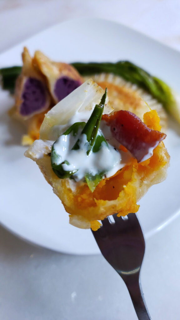 A closeup shot of the filling of these Vibrant 2-Color Sweet Potato Pierogi reveals a beautiful, harvest orange filling of traditional sweet potato. Another pierogi in the background is filled with purple sweet potato.
