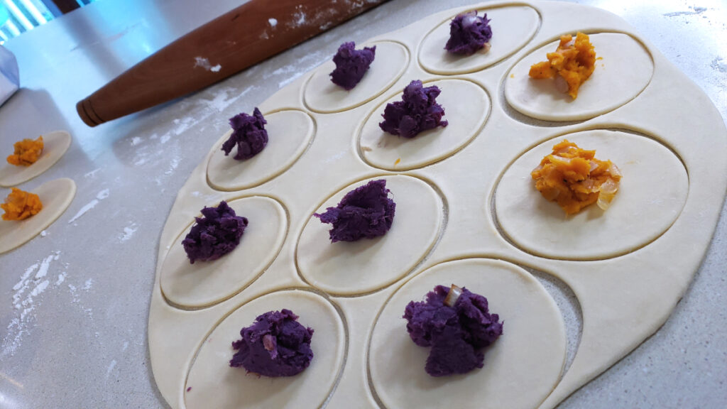 A closeup shot of the filling of these Vibrant 2-Color Sweet Potato Pierogi reveals a beautiful, harvest orange filling of traditional sweet potato. Another pierogi in the background is filled with purple sweet potato.