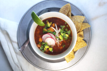 A beautiful bowl of Authentic Mexican Pozole Rojo in 45 Minutes sits at the center of a pewter serving plate that's adorned with crisp corn tortilla chips and a wedge of lime. Pork and hominy are simmered in a rich and vibrant red broth that is seasoned with achiote, cumin and cilantro. Fresh onions, cilantro, radishes and avocado adorn the top of the bowl.