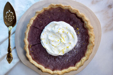 A serving platter and elegant golden pie knife frame this Stunning 8-Ingredient Purple Sweet Potato Pie reveals a vibrant purple color, and creamy texture. Topped with fresh whipped cream and lemon zest.