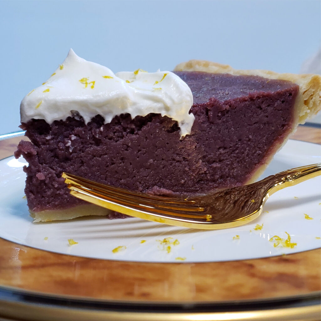 A slice of Stunning 8-Ingredient Purple Sweet Potato Pie reveals a vibrant purple color, and creamy texture. Topped with fresh whipped cream and lemon zest.
