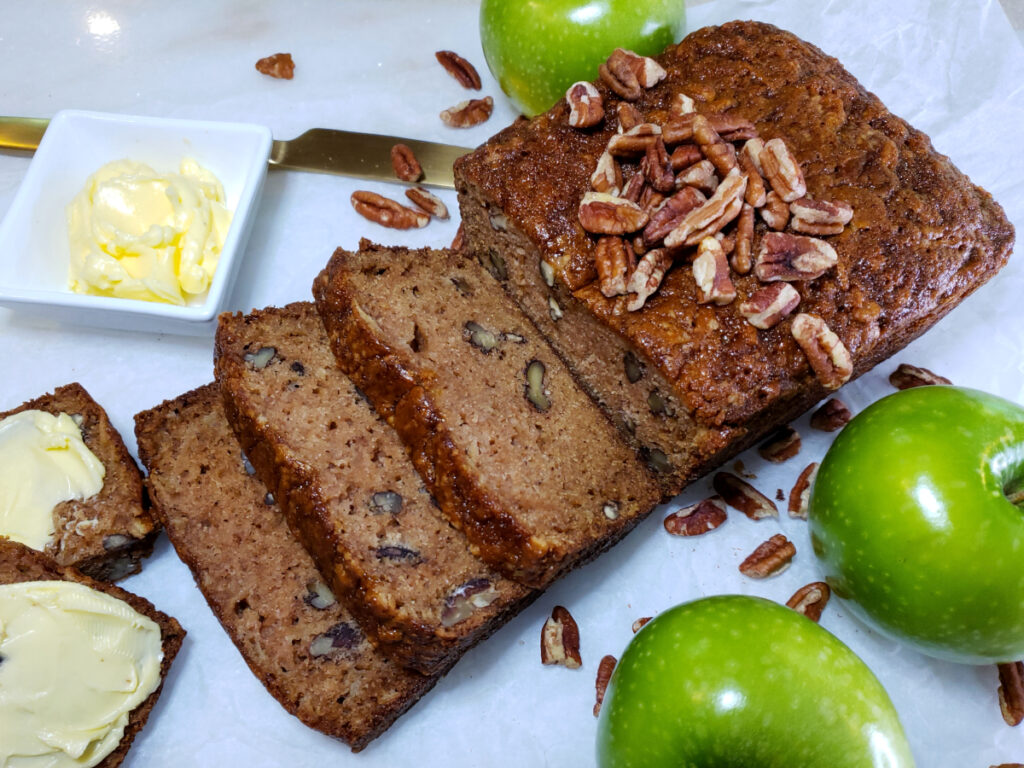 A fresh baked loaf of Easy Apple Quick Bread in 60 MInutes sits atop a cool marble cutting board. Three distinct slices reveal bits of shredded apple and pecans. Fresh Granny Smith apples and pecans frame the golden loaf. A small dish of whipped butter sits off to the left of the board. A small slice of the apple bread is generously topped with butter.