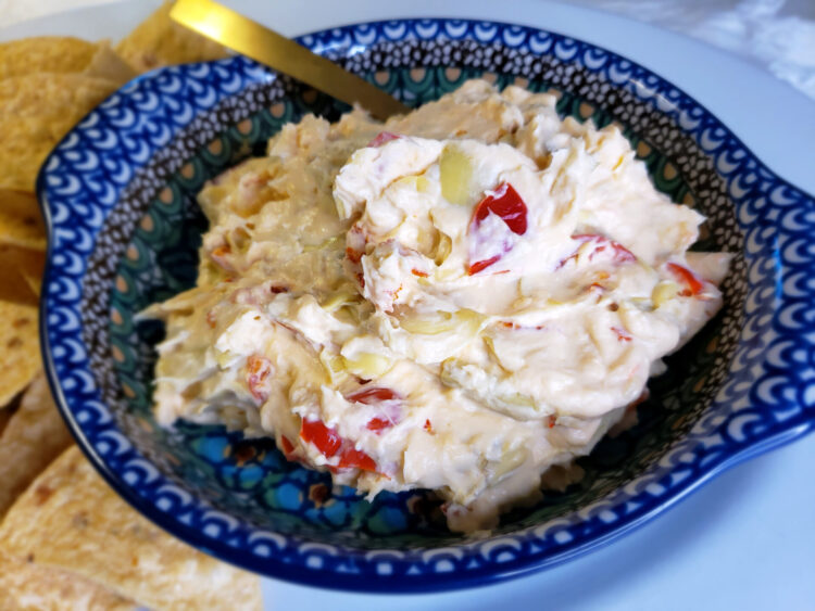 An oval platter is piled high with crisp tortilla chips. A side dish of this Easy Cheesy 10-Minute Peppadew Artichoke Dip sits off to the right side.
