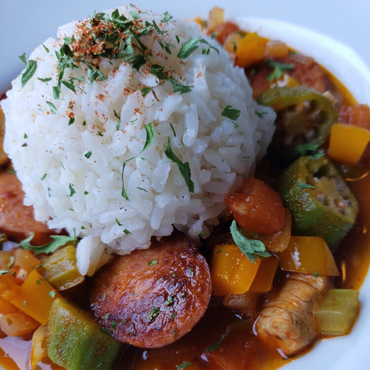A bowl of this Super Simple Chicken and Sausage Jambalaya in 1 Hour sits at the center of a tray. The spicy stew is packed with vegetables, chicken and sausage and topped with a generous scoop of white rice.