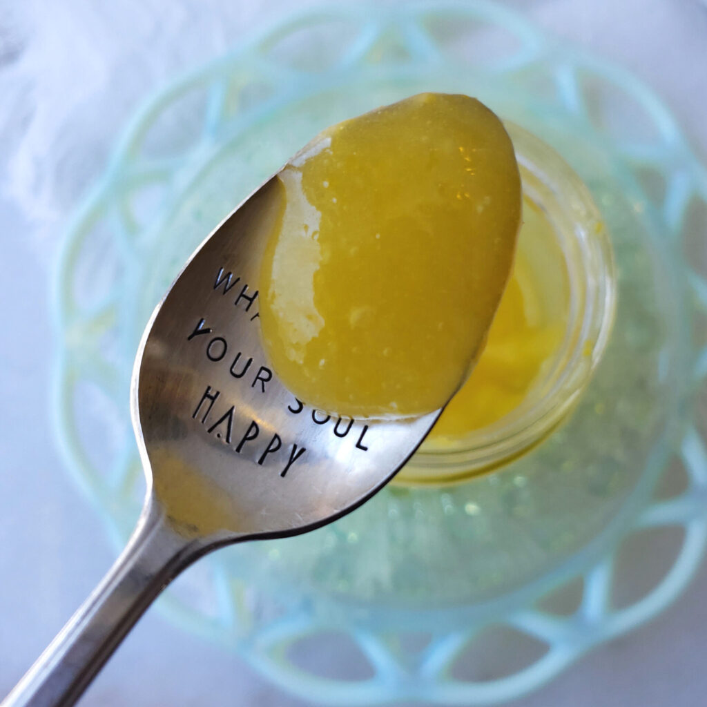 A spoonful of this Quick and Easy 4-Ingredient Lemon Curd hovers above a jar. The spoon is etched with the saying, "do something that makes your soul happy."